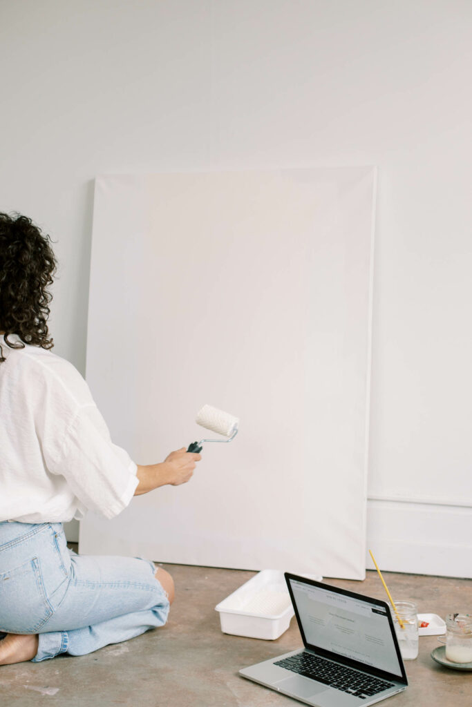 woman sitting on floor painting white onto white canvas, laptop resting on floor beside painting supplies